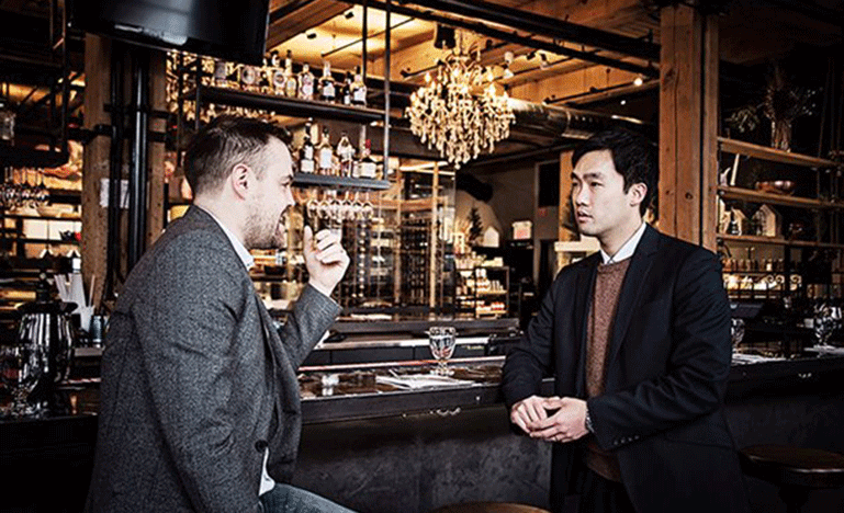 Two men wearing suits are at a Bar and are engaged in conversation.